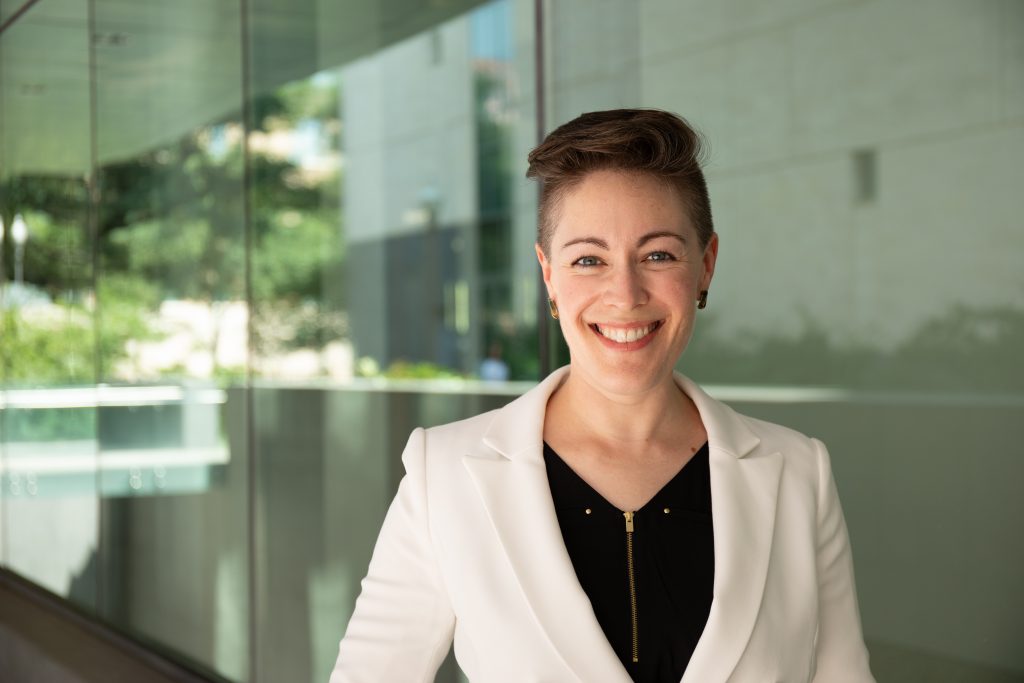 Woman in a blazer, smiling in front of a glass window.