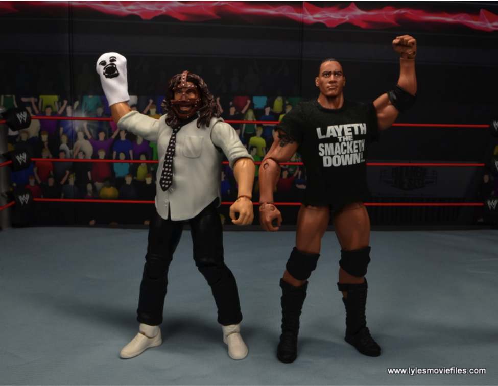 Two models of professional wrestlers Mankind and The Rock stand in a model wrestling ring, holding their arms raised, with a photo of an audience in the background. Mankind wears a sock puppet on one hand, and The Rock wears a t-shirt with the words "Layeth the Smacketh Down!" printed on it. 